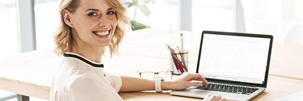 Lineas ICO - Mujer joven rubia de negocios sonriendo mientras se encuentra sentada en su escritorio con su portatil Lineas ICO - Mujer joven rubia de negocios sonriendo mientras se encuentra sentada en su escritorio con su portatil